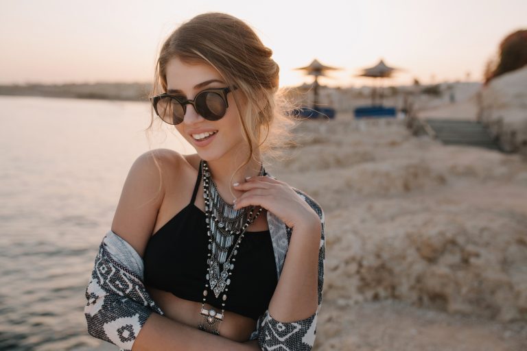 Une femme portant des lunettes de soleil et d'élégantes boucles d'oreilles se tient souriante près d'un plan d'eau avec un terrain rocheux en arrière-plan pendant le coucher du soleil.