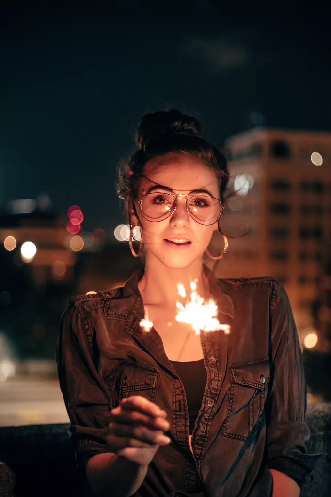 Une femme portant des lunettes et des créoles tient un cierge magique la nuit, avec des lumières floues de la ville en arrière-plan.