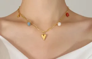 Gros plan sur une personne portant un collier en chaîne en or avec un pendentif en forme de cœur et des breloques multicolores, vêtue d'un haut blanc à épaules nues.