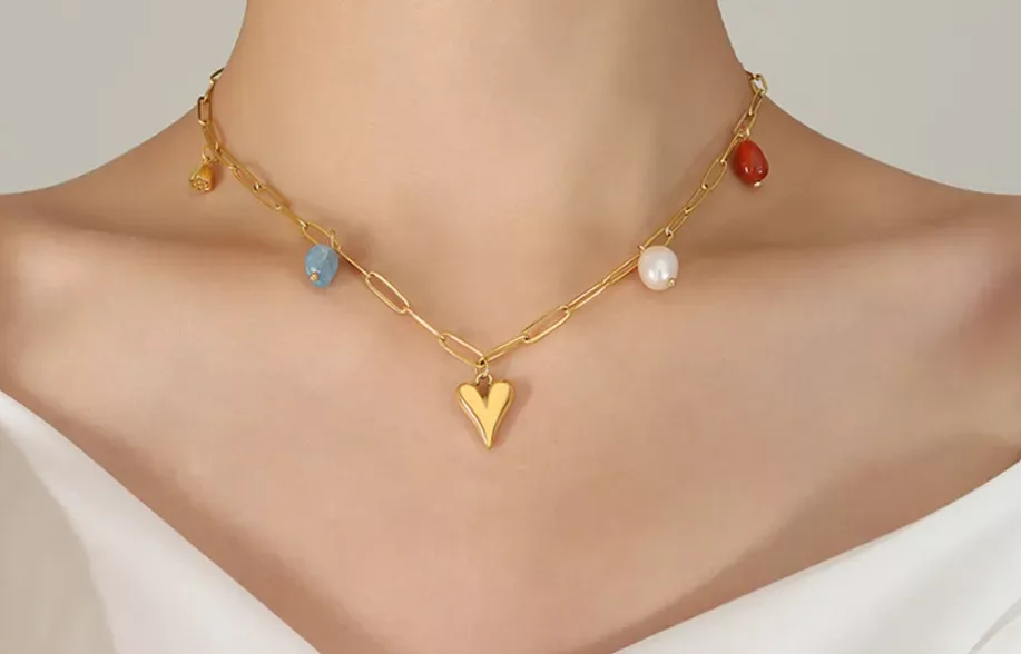 Gros plan sur une personne portant un collier en chaîne en or avec un pendentif en forme de cœur et des breloques multicolores, vêtue d'un haut blanc à épaules nues.