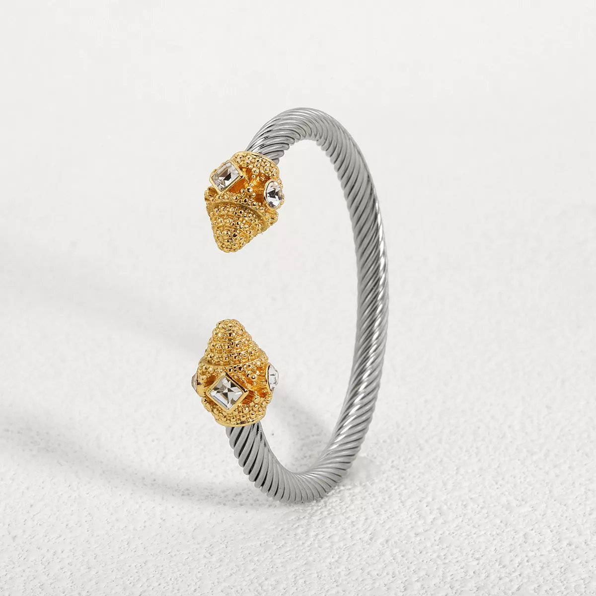 Un bracelet manchette en câble torsadé en argent avec des embouts texturés en or, chacun orné d'une pierre précieuse transparente, présenté sur une surface blanche.
