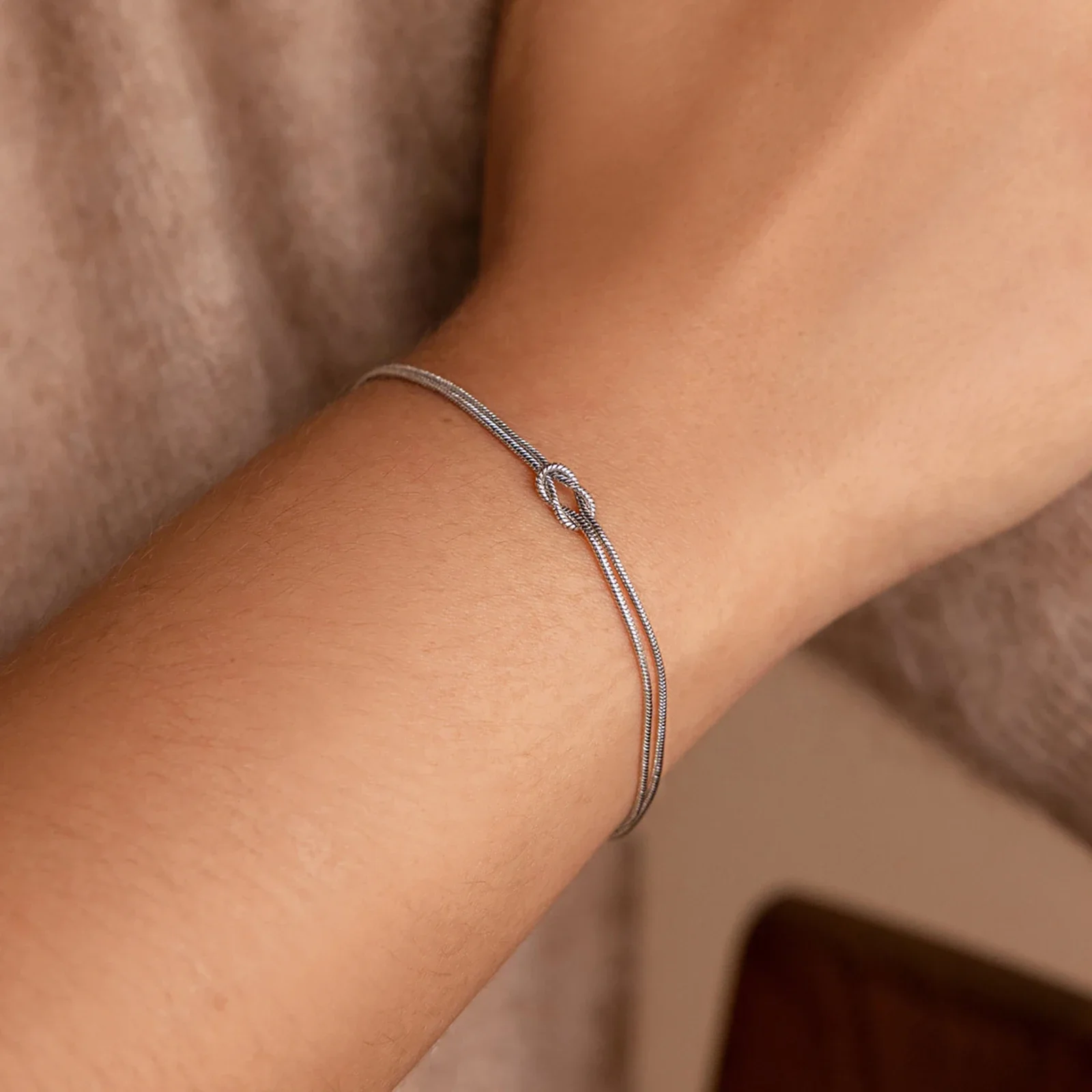 Gros plan sur un poignet portant un fin bracelet en argent avec un petit nœud, sur un fond neutre.