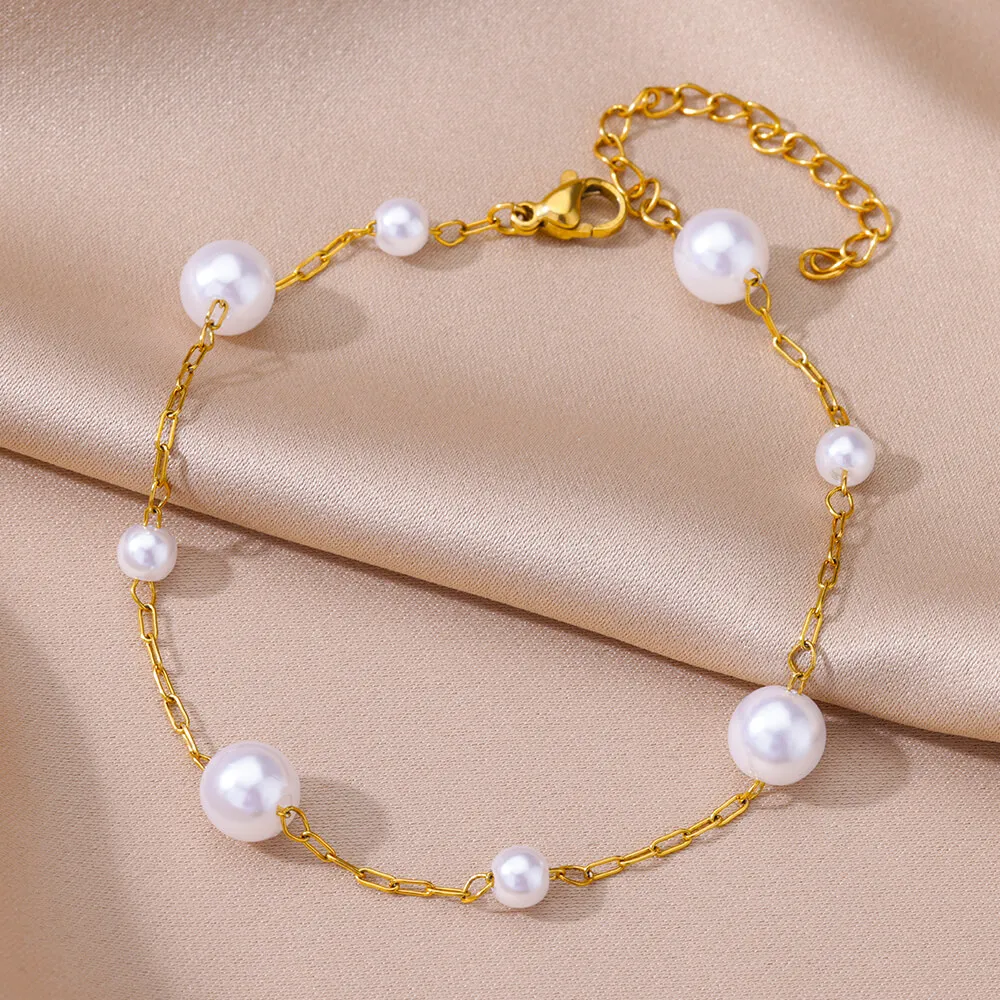 Un bracelet en chaîne d'or avec sept perles blanches de tailles différentes, présentées sur un fond de tissu satiné beige.