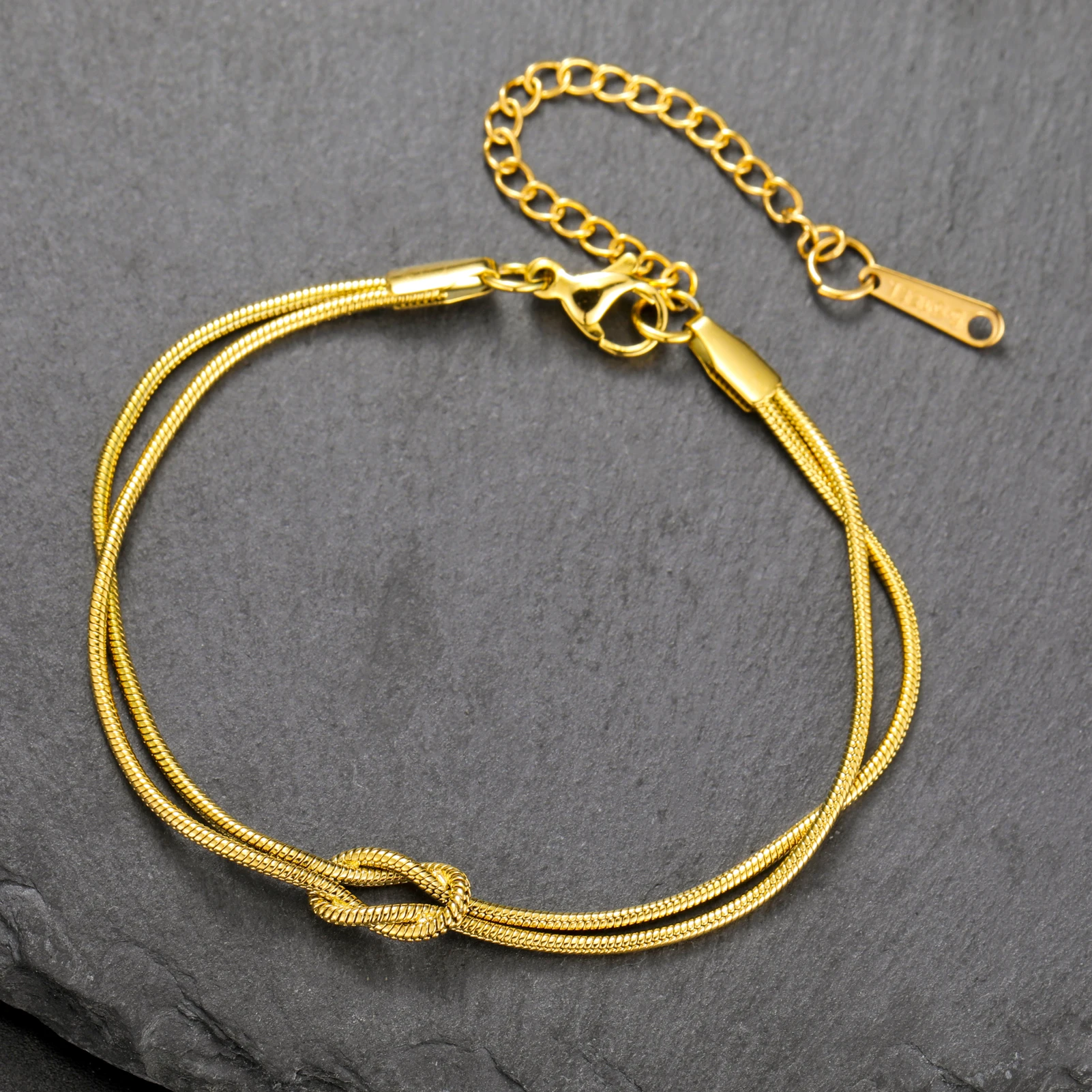 Bracelet en métal doré avec un détail de nœud et un fermoir en chaîne réglable, présenté sur une surface de pierre texturée foncée.