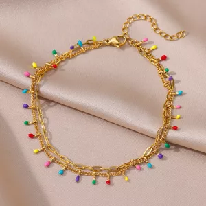 Un bracelet de cheville en chaîne dorée, orné de petites perles colorées réparties uniformément sur un fond de tissu satiné beige clair.