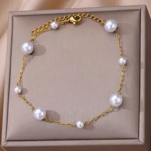 Un bracelet en chaîne en or avec six perles blanches régulièrement espacées, présentées sur une surface beige coussinée.