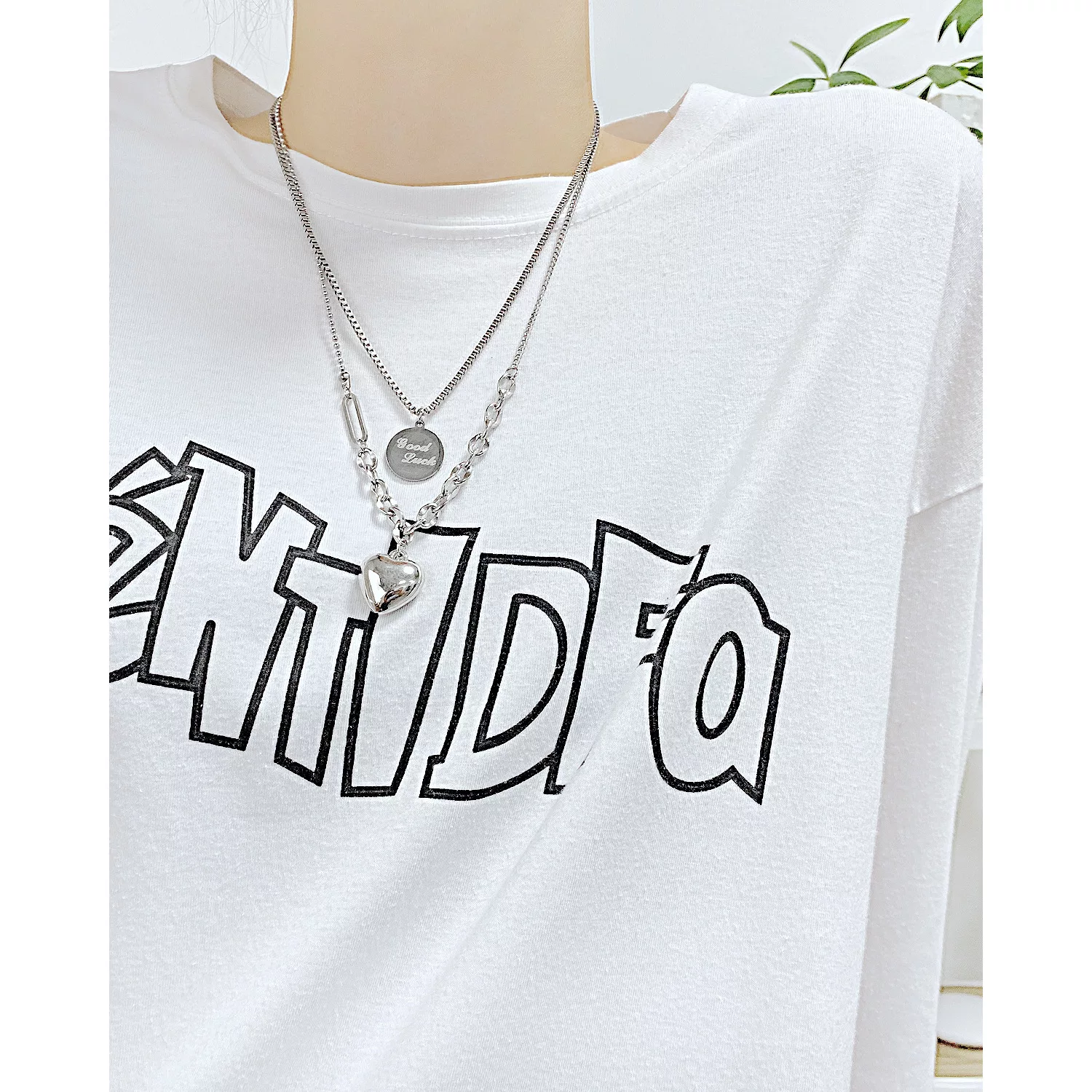 Une personne portant une chemise blanche avec un texte noir en gras et deux colliers en argent, l'un avec un pendentif rond et l'autre avec un pendentif en forme de cœur.