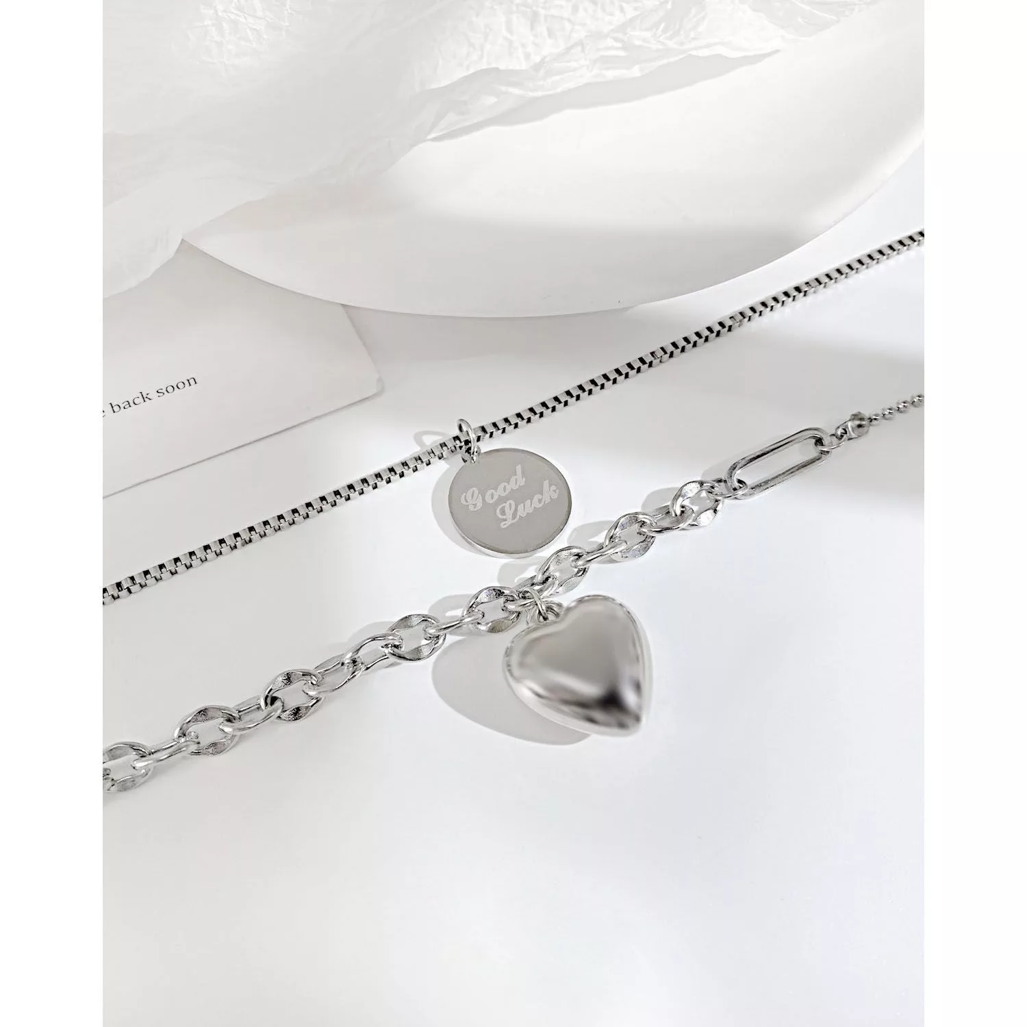 Deux colliers en chaîne en argent, l'un avec un pendentif rond "Good Luck" et l'autre avec un pendentif en forme de cœur gonflé, présentés sur une surface blanche.