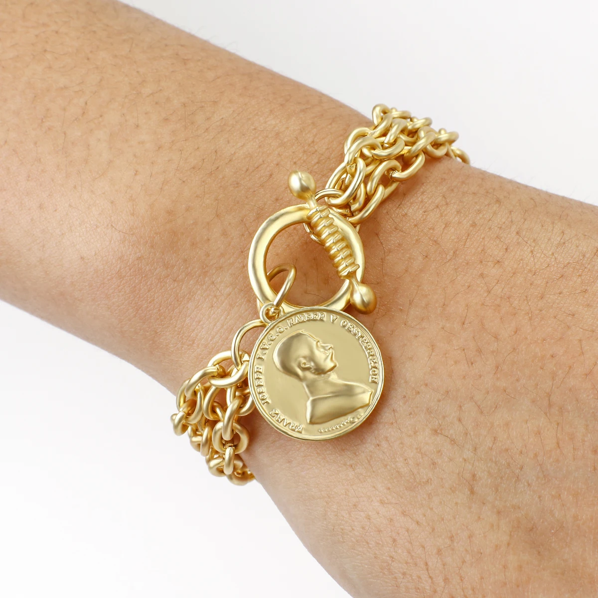 Bracelet en chaîne en or avec un médaillon rond représentant le profil d'un visage d'homme, porté au poignet d'une personne.