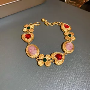 Un bracelet en or orné de pierres rouges en forme de cœur, de fleurs en pierres vertes et de breloques représentant des portraits roses de style camée est présenté sur un fond gris.