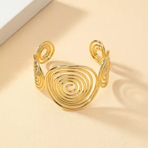 Un bracelet manchette en or, orné de motifs en spirale réalisés en fil métallique, est présenté sur un fond beige.