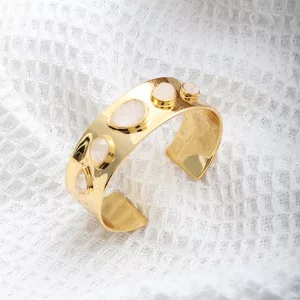 Un bracelet manchette en or, orné d'incrustations de pierres blanches ovales et rondes, est présenté sur un fond en tissu blanc texturé.