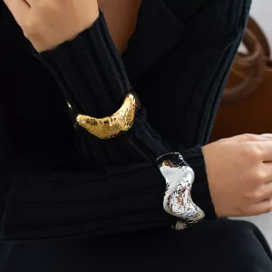 Une personne portant un haut noir à manches longues en maille côtelée, avec un large bracelet texturé doré à un poignet et un bracelet argenté similaire à l'autre.