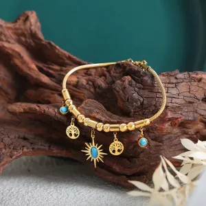 Un bracelet en or orné de pierres bleues et de breloques pendantes en forme de soleil et d'arbre, présenté sur un morceau de bois texturé, avec un feuillage blanc décoratif à proximité.