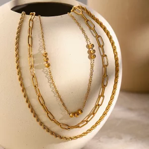 Trois colliers en or, aux styles de chaînes variés, sont disposés sur un vase en céramique beige, sur fond neutre.