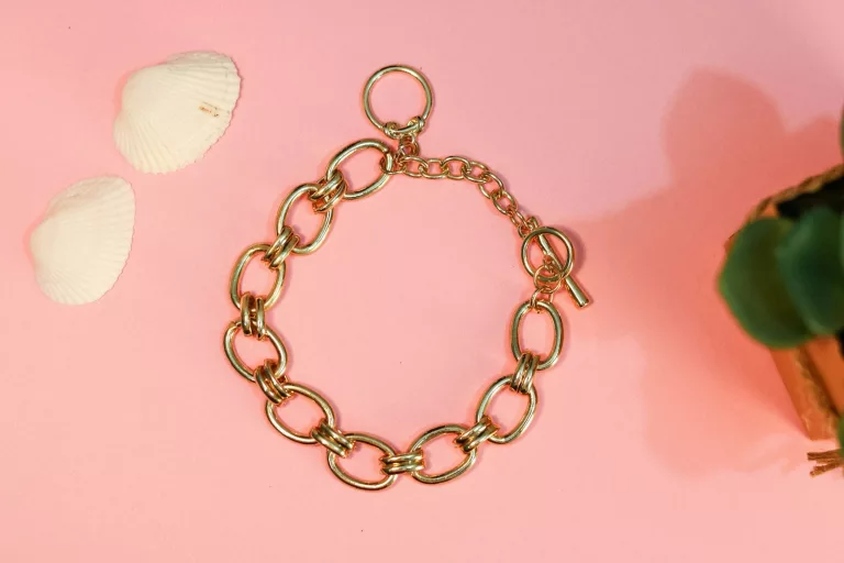 Bracelet en chaîne dorée composé de gros maillons ovales et muni d'un fermoir à bascule, présenté sur un fond rose, aux côtés de deux coquillages blancs et d'un fragment de plante verte.